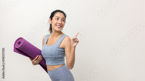 Young beautiful asian woman with sportswear holding yoga mat on pointing to blank space isolated background. Portrait sporty woman standing pose exercise workout in studio. Training yoga lifestyle.