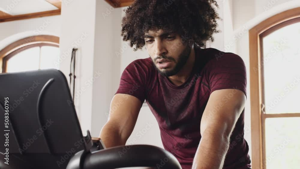 Focused young adult Middle Eastern male cycling on indoor fitness bike, determined and tired in an indoor fitness gym.