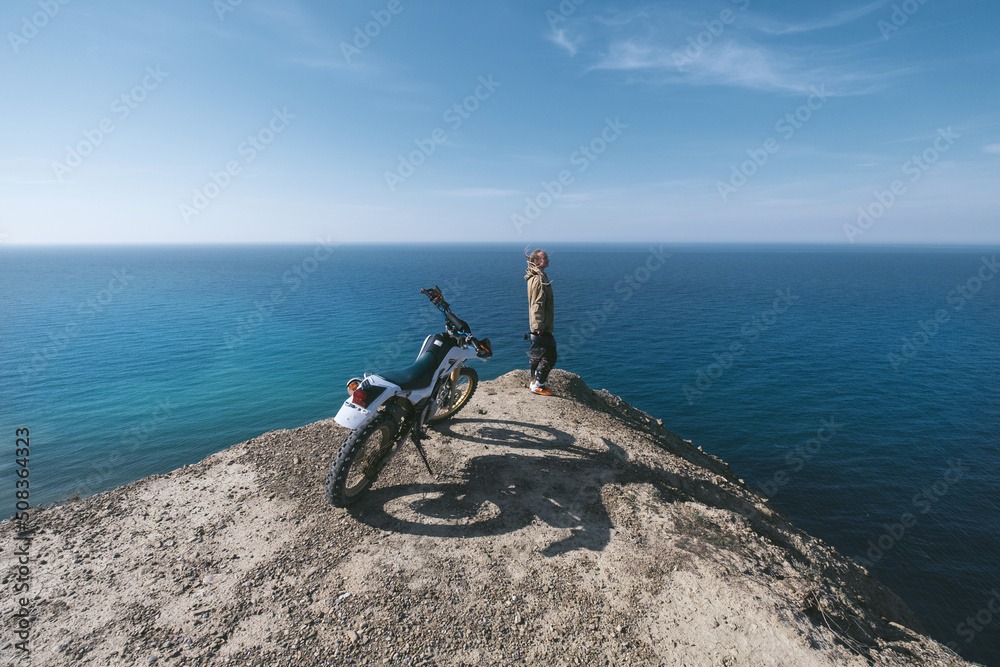 Female rider relaxing in enduro offroad motorcycle travel on mountain top, beautiful sea shore and mountains landscape on background