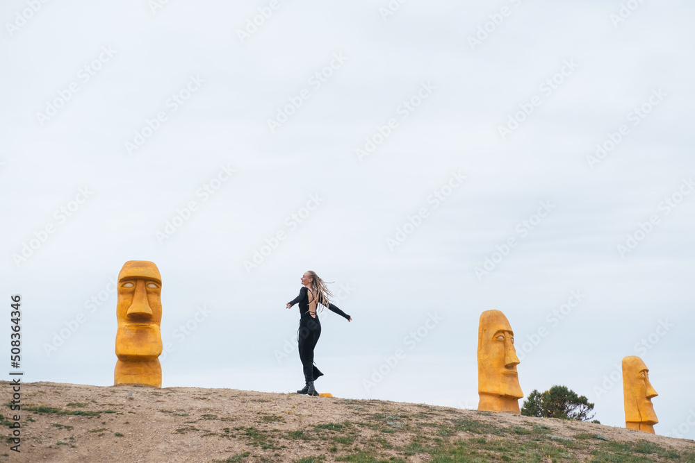 Obraz premium Woman wearing dreadlocks and long black dress walking near wooden statues like on easter island by the sea shore