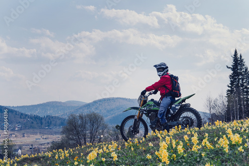 Female wearing helmet sitting on dirt motorcycle on field with yellow flowers and pine forest