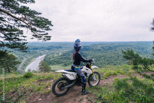 female biker wearing helmet and pink dreadlocks sitting on her dirt motorcycle on mountain top above Beautiful view