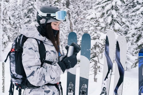 Woman skier resting, standing near freeride skis opening can of drink. Deep winter forest, trees covered snow photo