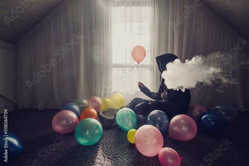 Young man wearing hoodie sitting near the window in  empty room with balloons and smoke