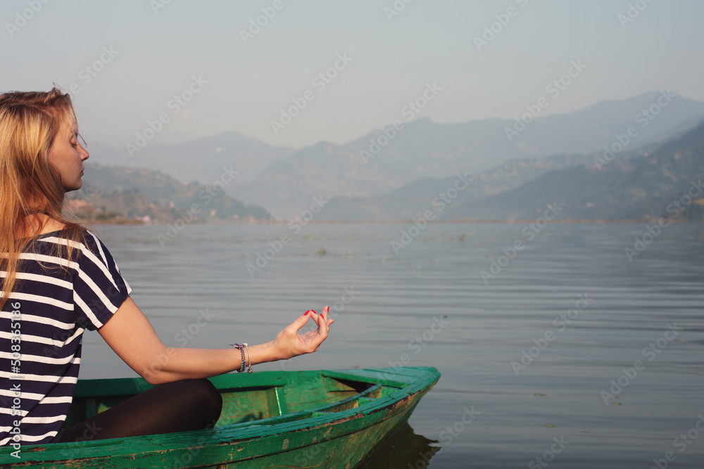 young female sitting in meditating pose on sunny summer day in wooden boat on fabulous mountain lake. Meditation outdoor