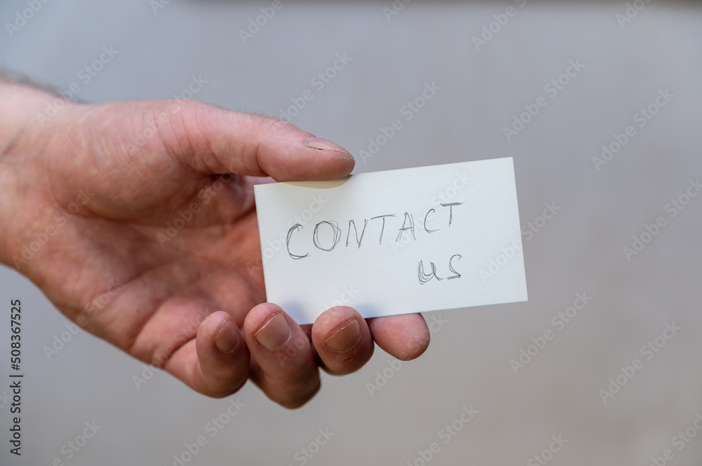 Contact us. Words written in jagged letters. A man's hand holds a white paper rectangle with text against a gray background. Contact us. Words written in jagged letters. A man's hand holds a white paper rectangle with text against a gray background.