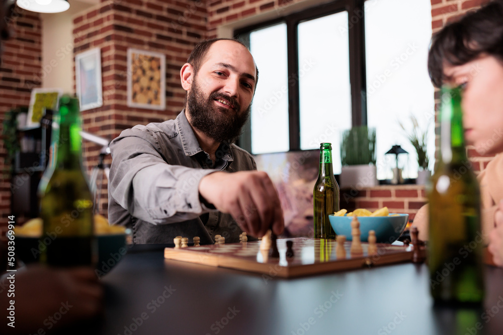 Smiling man moving chess piece on board while sitting at table with close friends. Multiethnic people in living room at home playing board games while consuming snacks and beverages.