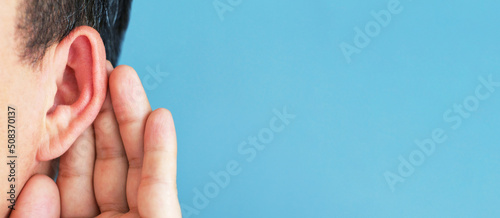 Man put his hand to his ear and listens carefully on blue background copy space.