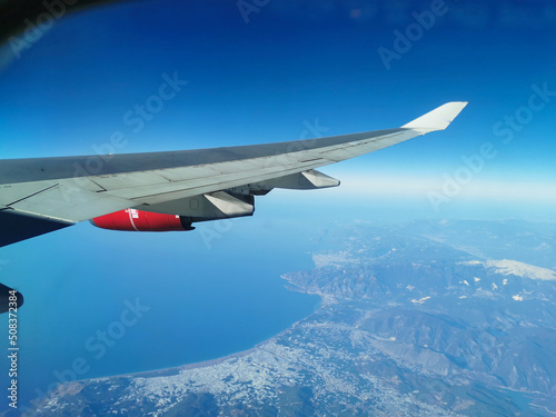 View from the porthole on the wing of an airplane with a red aircraft engine, in the morning over beautiful mountains with snowy peaks and the shore of the Mediterranean Sea.