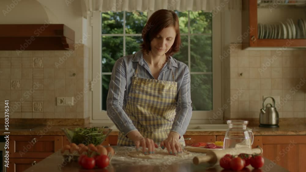 30s woman in apron placing grated cheese on pizza dough standing in the kitchen. Female chef cooking Italian homemade pizza. 30s woman in apron placing grated cheese on pizza dough standing in the kitchen. Female chef cooking Italian homemade pizza.