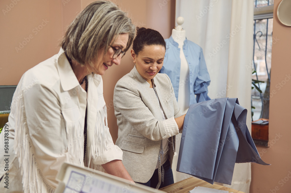 Portrait of a middle-aged Hispanic fashion designer in a stylish jacket holding blue fabric to create a new collection of women's dress shirts in a fashion design workshop