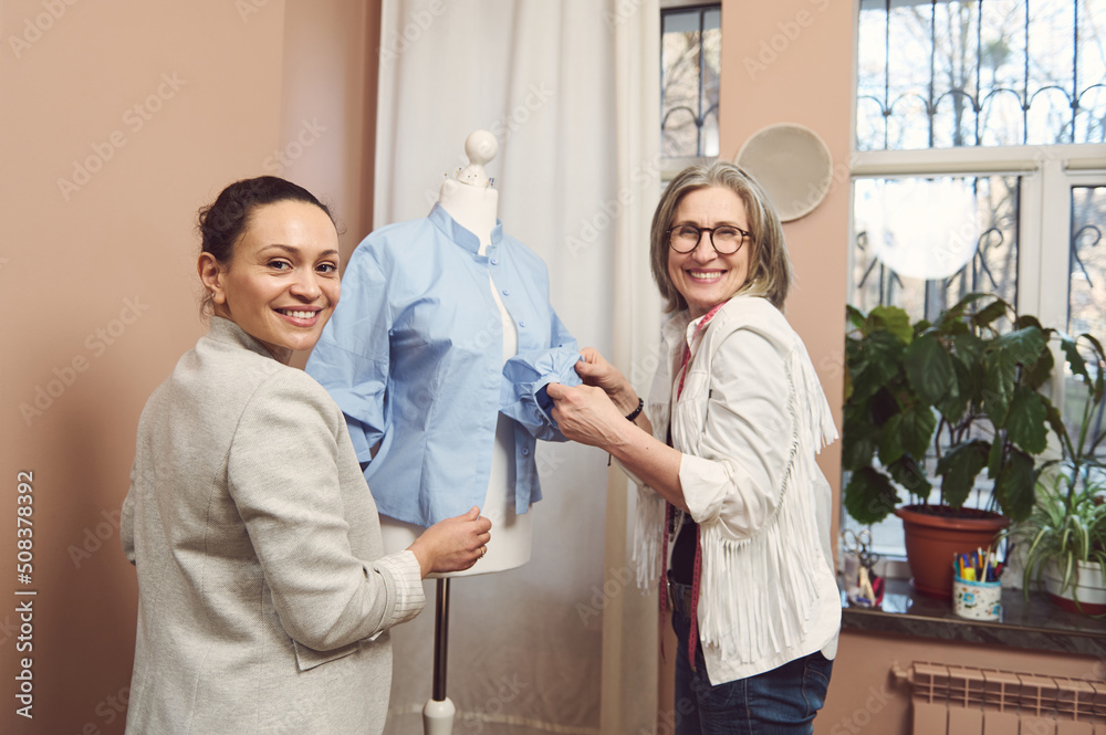 Beautiful multi-ethnic female seamstresses working with a blue shirt on a dressmaker's model, improving details of new collection in a fashion design atelier. Fashion designer business start-up Beautiful multi-ethnic female seamstresses working with a blue shirt on a dressmaker's model, improving details of new collection in a fashion design atelier. Fashion designer business start-up