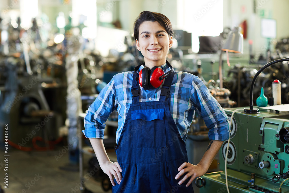 Cheerful successful lady machinist with ear protectors on neck standing in workshop of industrial plant and holding hands on hips while looking at camera Cheerful successful lady machinist with ear protectors on neck standing in workshop of industrial plant and holding hands on hips while looking at camera