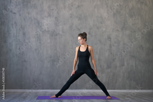 Attractive young woman doing yoga stretching yoga online at home against gray wall in black sportswear. Self-isolation is useful entertainment and education on Internet. The concept healthy lifestyle.