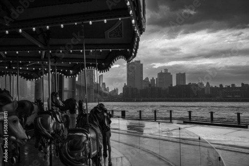 Fotografie manège de chevaux de bois ancien au premier plan avec en arrière plan Manhattan et la skyline sous l'orage à New York