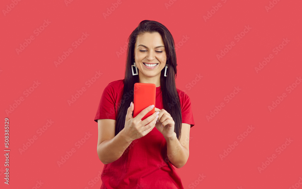 Happy beautiful brunette woman using modern mobile phone. Pretty young lady standing on red background, holding her smartphone in red phone case, downloading new apps, reading new messages and smiling Happy beautiful brunette woman using modern mobile phone. Pretty young lady standing on red background, holding her smartphone in red phone case, downloading new apps, reading new messages and smiling