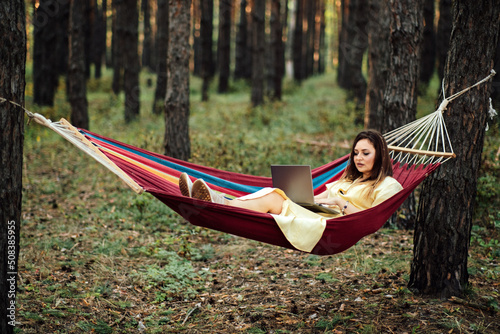 Young happy successful woman freelancer with laptop relaxing in the hammock on summer Pine forest on sunny day. Female entrepreneur working with laptop in hammock and camping in nature.