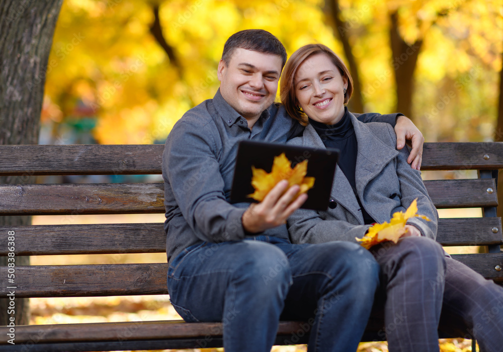 Fototapeta premium a woman and a man are sitting on a bench in an autumn park, they have a pad and a bouquet of yellow maple leaves, a bright sunny day