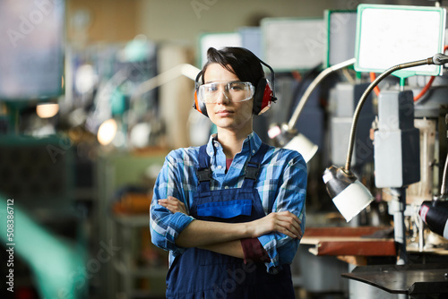 Serious gritty woman in safety goggles and ear protectors standing at industrial shop and crossing arms on chest while looking at camera