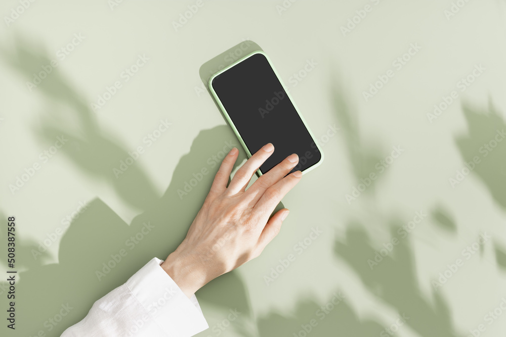 Woman hold phone with black blank screen, minimal flat lay on olive green background. Smartphone mock up. Digital online summer lifestyle composition with leaves shadows and sunlight
