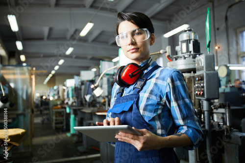 Content attractive masculine lady worker in safety goggles wearing ear protectors on neck working with online data on table and looking at camera