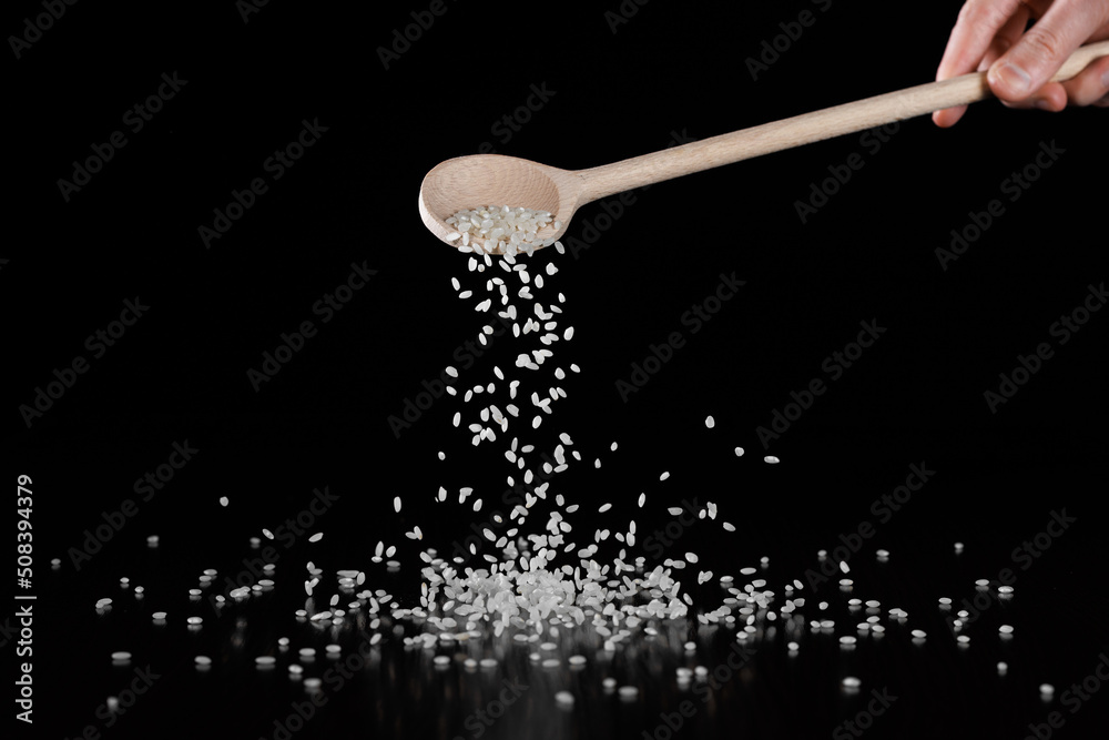 White round rice is poured from a wooden spoon onto the table. Selective focus. Black background.