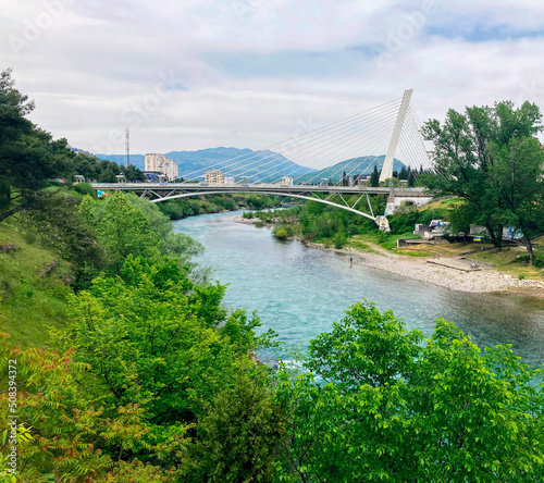 Park area overlooking a mountain river and a large white suspension bridge. The river is clear with turquoise water. A lot of greenery grows along the river.