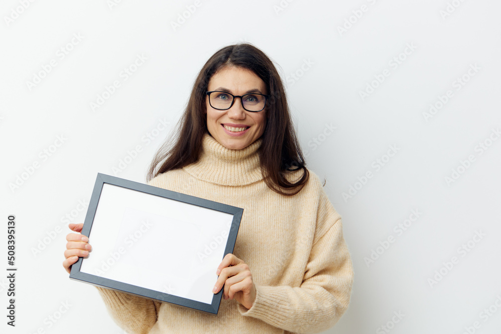 neat woman in a beige turtleneck sweater and black-rimmed glasses, standing on a light background holding a white frame with a black frame in her hands. Horizontal photo for advertising text neat woman in a beige turtleneck sweater and black-rimmed glasses, standing on a light background holding a white frame with a black frame in her hands. Horizontal photo for advertising text