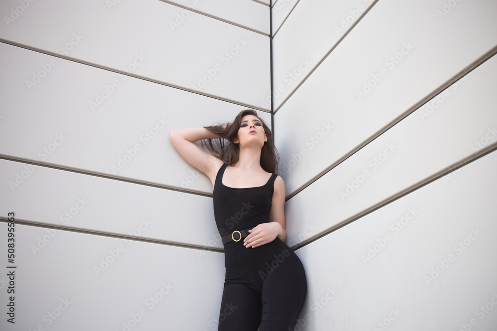 Young woman in black top and leggings leaning on white wall and looking at camera