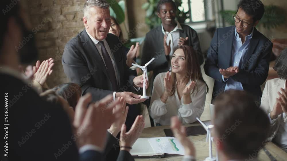 young coworkers clapping hands to business boss holding a windmill turbine in hand - celebration of the success of the project and teamwork