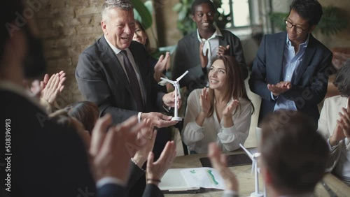 young coworkers clapping hands to business boss holding a windmill turbine in hand - celebration of the success of the project and teamwork photo