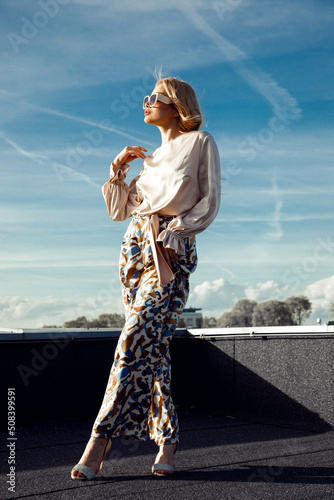 Young woman with long fair hair wearing silk blouse  printed trousers  sunglasses  standing with crossed legs on roof.
