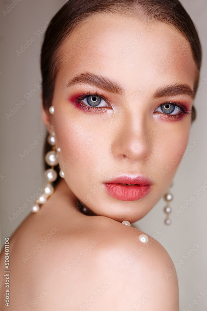Headshot of pretty young woman with makeup and pearls on bare shoulders looking at camera on gray background 