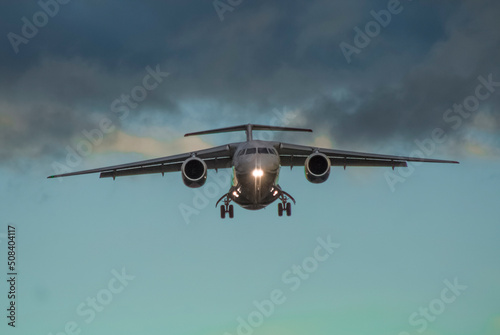 A civilian jet comes in to land at an airport at sunset with landing gear extended and landing lights on, front photo