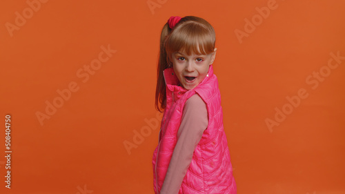 Funny joyful sincere little girl 6 years old in vest making playful silly facial expressions and grimacing  fooling around  turning  smiling. Young toddler child isolated alone on orange background