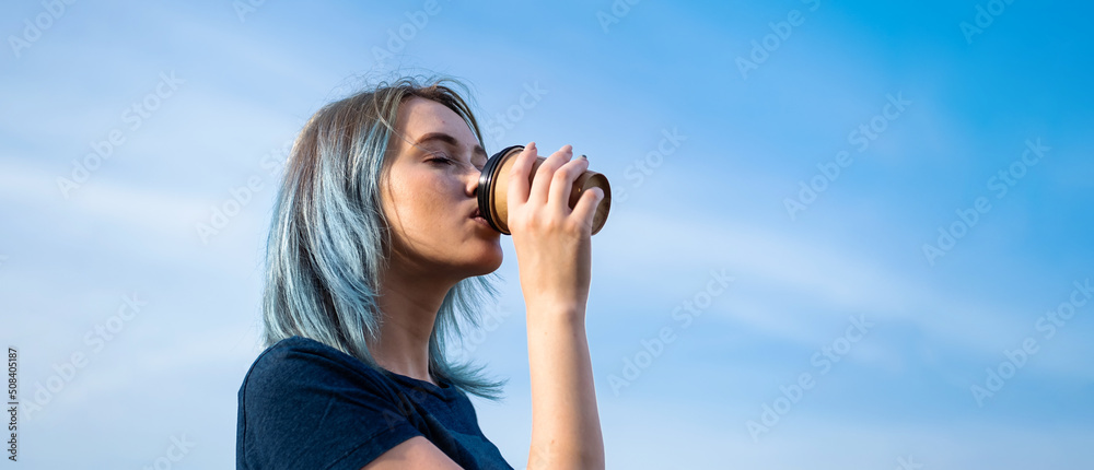 Blue haired woman drinks take away coffee against blue sky.