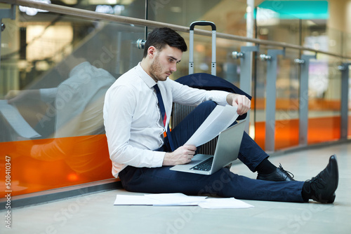Serious concentrated bearded businessman in formalwear sitting on floor in waiting area of airport and working with papers while using laptop