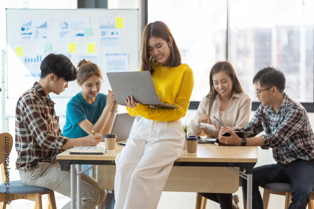 Fototapeta premium Portrait of happy smiling young woman holding a laptop in office meeting room looking at camera while colleagues are meeting in the background.
