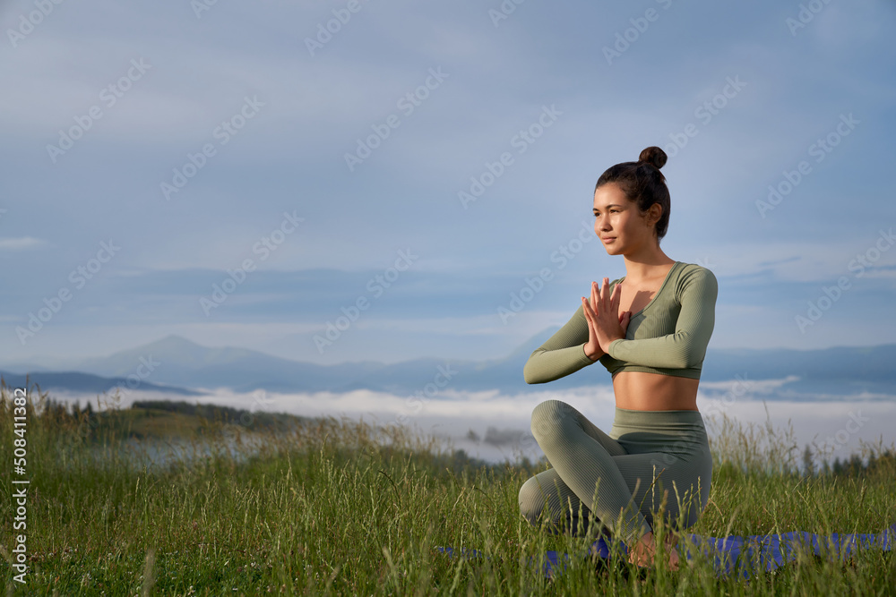 Sporty dark haired woman in activewear meditating with namaste gesture among green mountains. Young lady enjoying calmness and harmony on wild nature. Sporty dark haired woman in activewear meditating with namaste gesture among green mountains. Young lady enjoying calmness and harmony on wild nature.