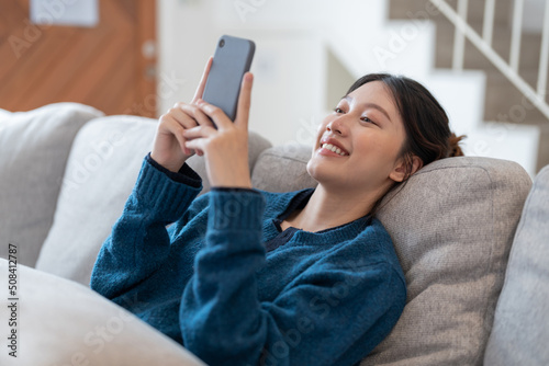 Fotografie Happy young asian woman lying on sofa at living room and using cell phone at hom
