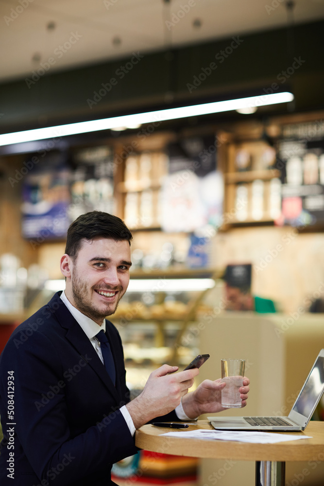 Portrait of cheerful confident young dealing manager in suit sitting at table and texting sms on smartphone while drinking water and waiting for company representative Portrait of cheerful confident young dealing manager in suit sitting at table and texting sms on smartphone while drinking water and waiting for company representative