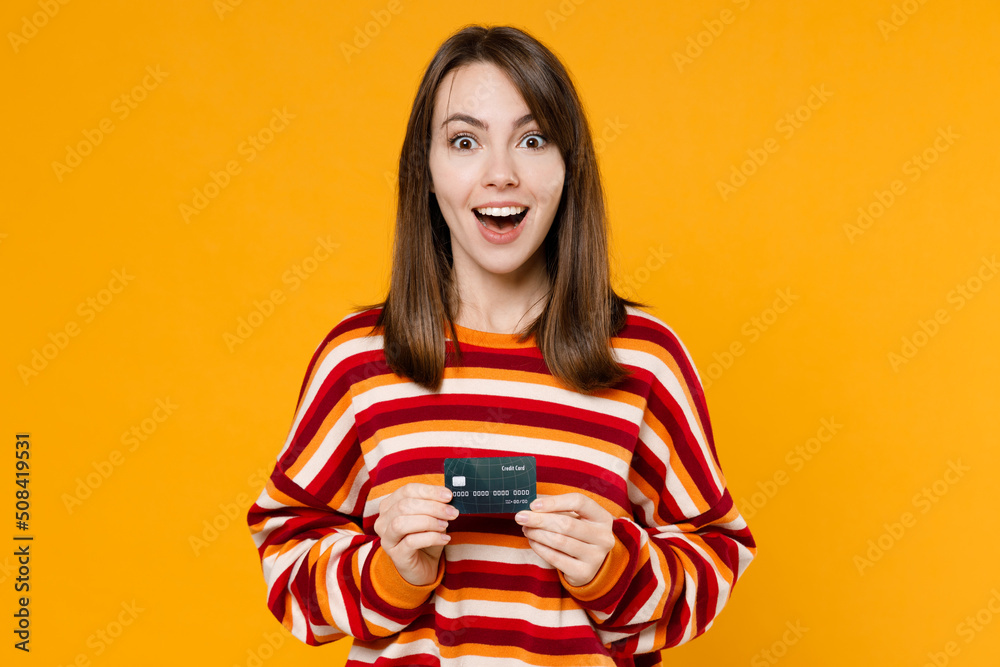 Young happy shocked amazed impressed excited caucasian woman 20s in red striped sweatshirt hold in hand credit bank card isolated on plain yellow background studio portrait. People lifestyle concept. Young happy shocked amazed impressed excited caucasian woman 20s in red striped sweatshirt hold in hand credit bank card isolated on plain yellow background studio portrait. People lifestyle concept.