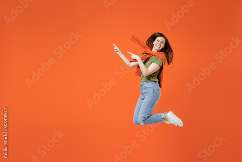 Full body side view young happy woman in khaki t-shirt tied sweater on shoulders look camera point index finger aside on workspace area isolated on plain orange background. People lifestyle concept.