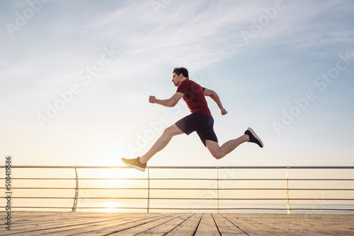 Side view full body young strong sporty athletic fit sportsman man in sports clothes run jump high warm up training at sunrise sun dawn over sea beach outdoor on pier seaside in summer day morning