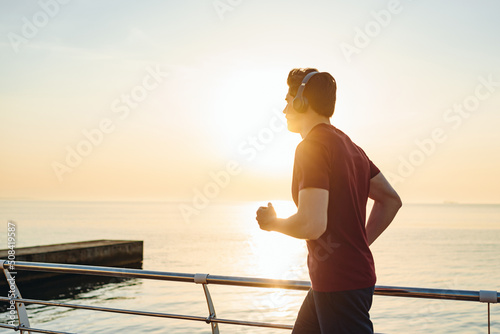 Side view sunlit young strong sporty athletic fit sportsman man wearing sports clothes heapdhones listen music warm up training at sunrise sun dawn over sea beach outdoor on pier seaside in morning.