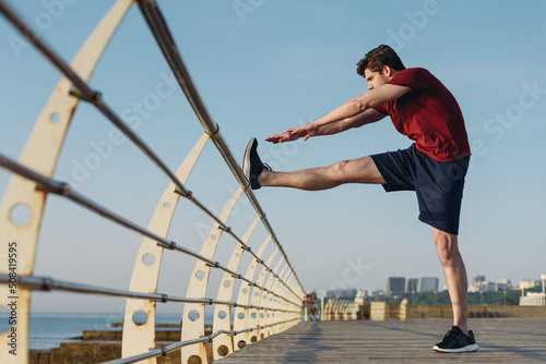 Full size side view young strong sporty fit sportsman man 20s in sports clothes doing stretch exercise for legs warm up training at sunrise sun dawn over sea beach outdoor on pier seaside in morning © ViDi Studio