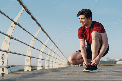 Full body strong sporty athletic toned fit sportsman man in sports clothes sit laces up sneakers shoes warm up train at sunrise sun dawn over sea beach outdoor on pier seaside in summer day morning © ViDi Studio