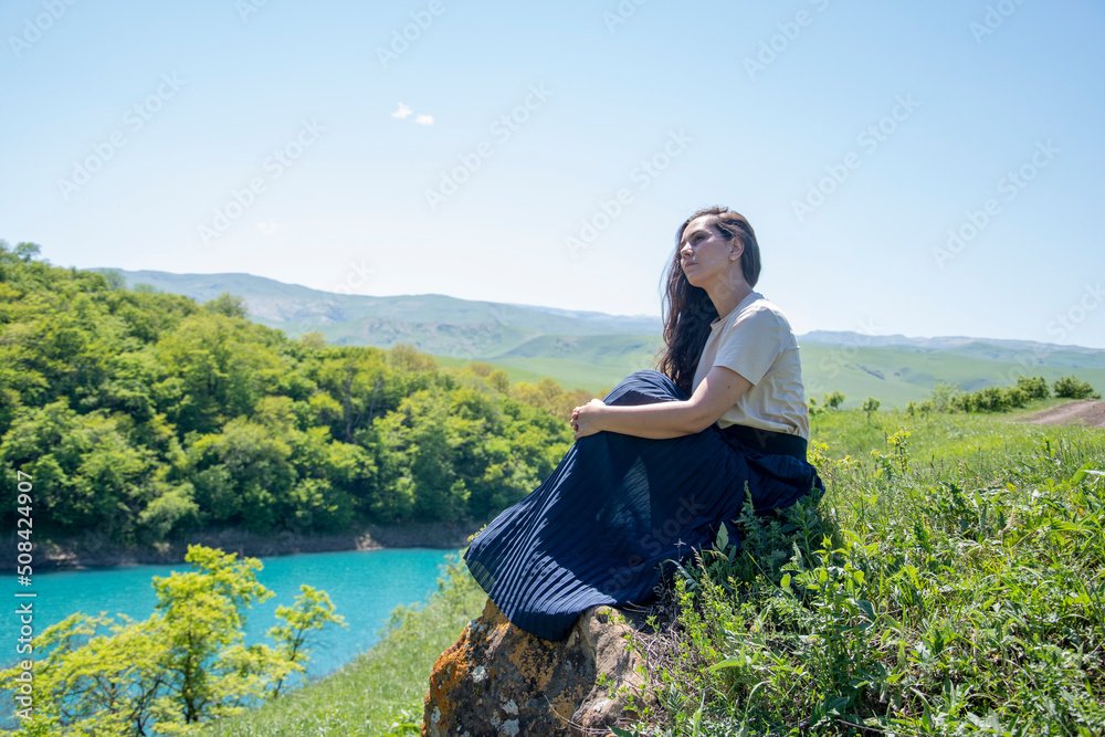 A girl in a dress sits on the shore of a blue lake in summer