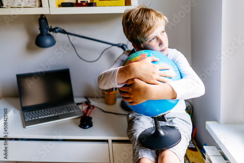 Smart sad child holding globe sitting on workplase in his room at home. The boy dreams and bored at home by himself. photo