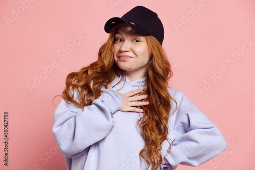 Stylish young redhead girl, student in hoodie and cap isolated on pink studio background. Fashion, human emotions, facial expression concept. Trendy colors